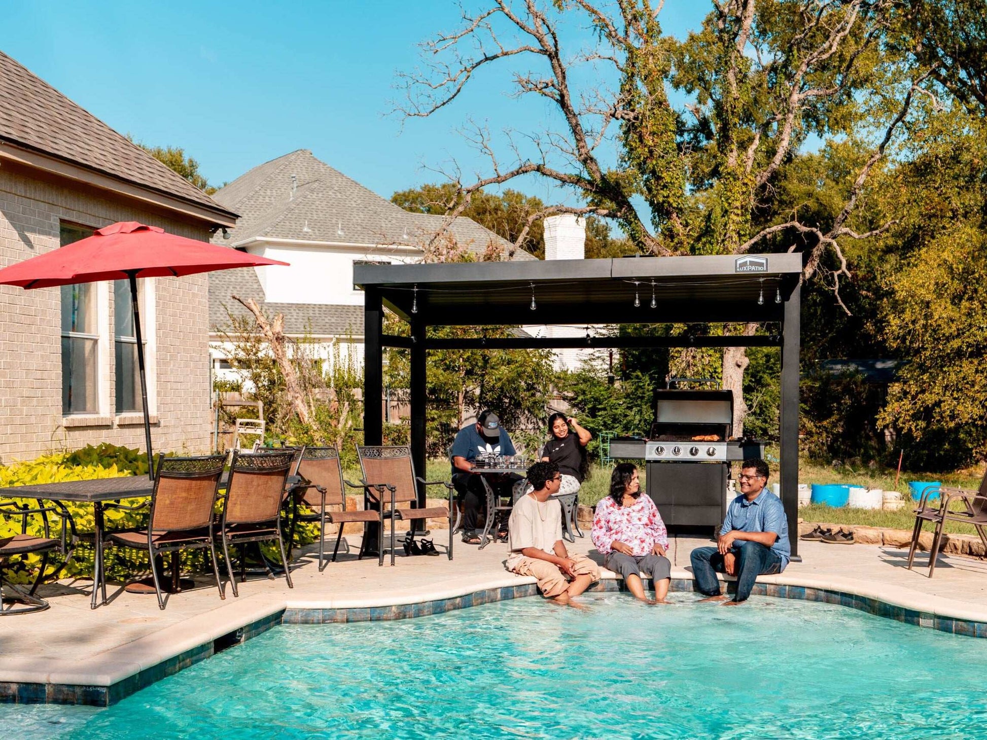 A family enjoying a sunny day by a swimming pool, with some sitting at the edge and others under a black pergola.