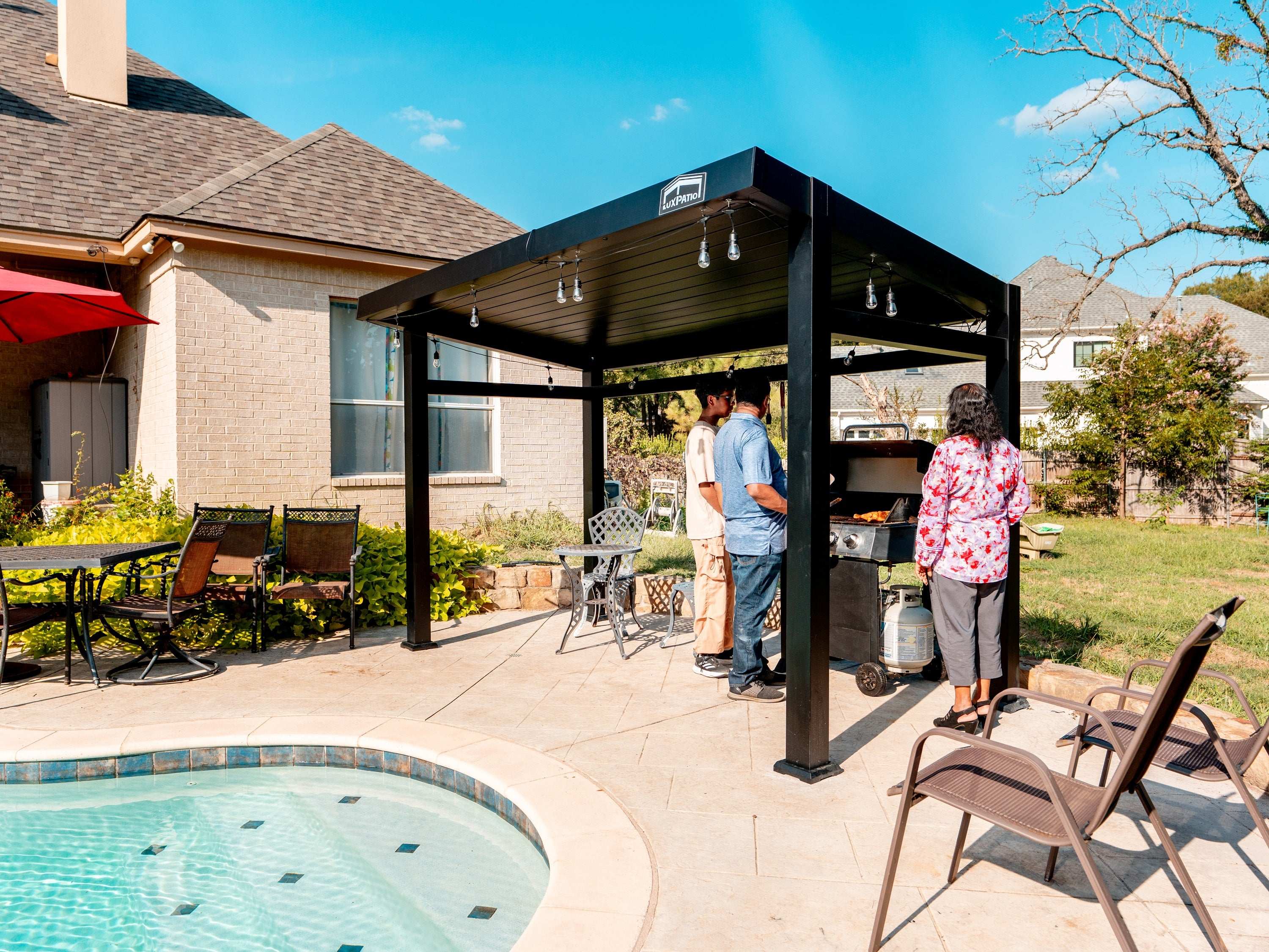 People gathered under a black pergola with string lights, next to a pool and a grill on a backyard patio.