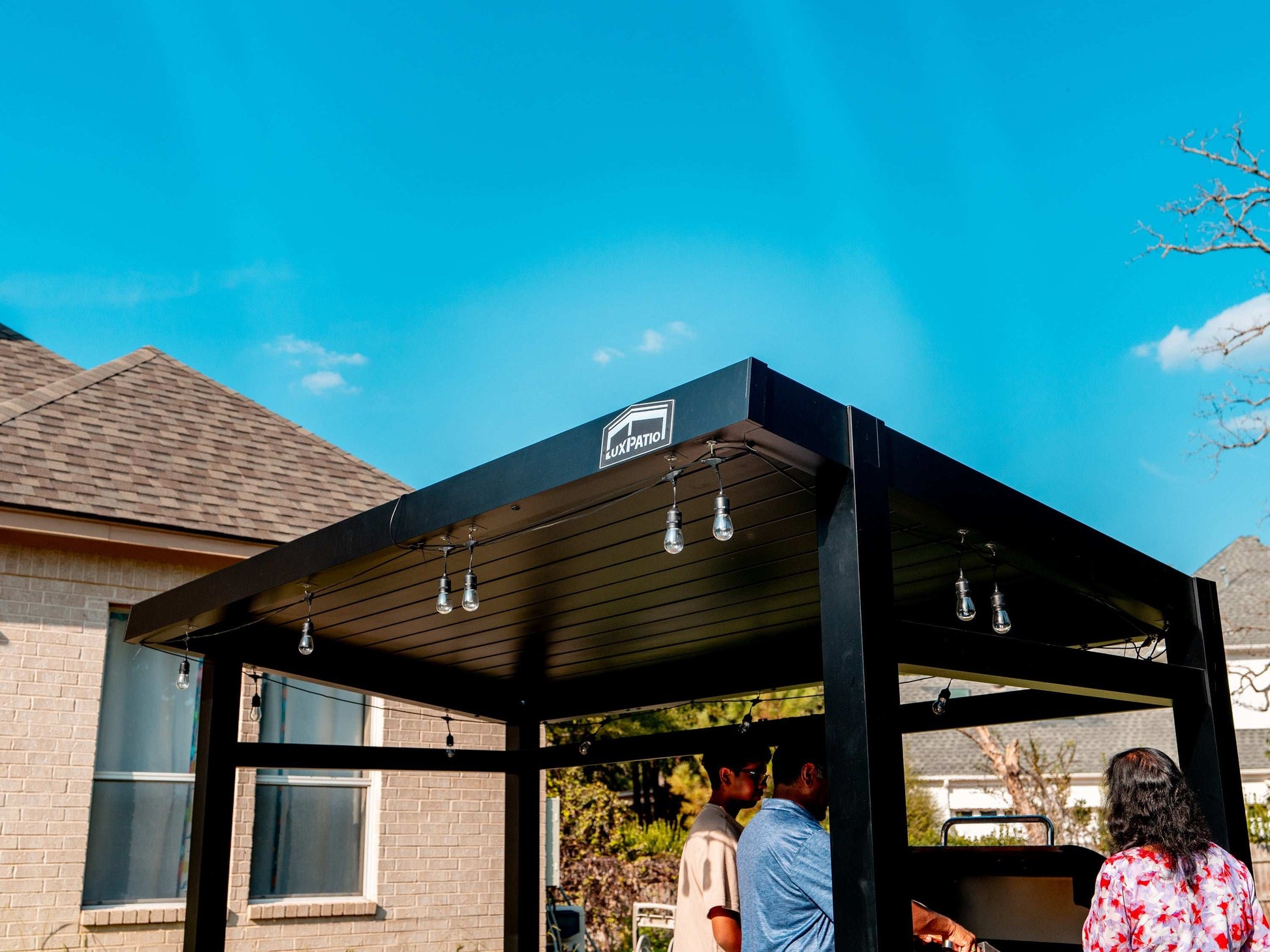 A black pergola with a solid roof and string lights over a patio, with people standing underneath and a brick house nearby.