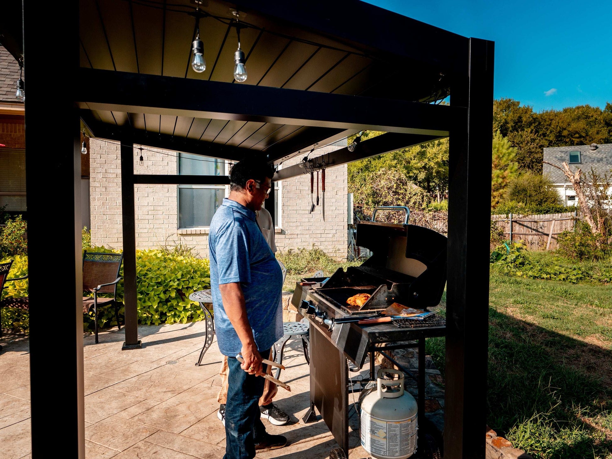 A man grilling under a black pergola on a patio in a sunny backyard with a brick house in the background.