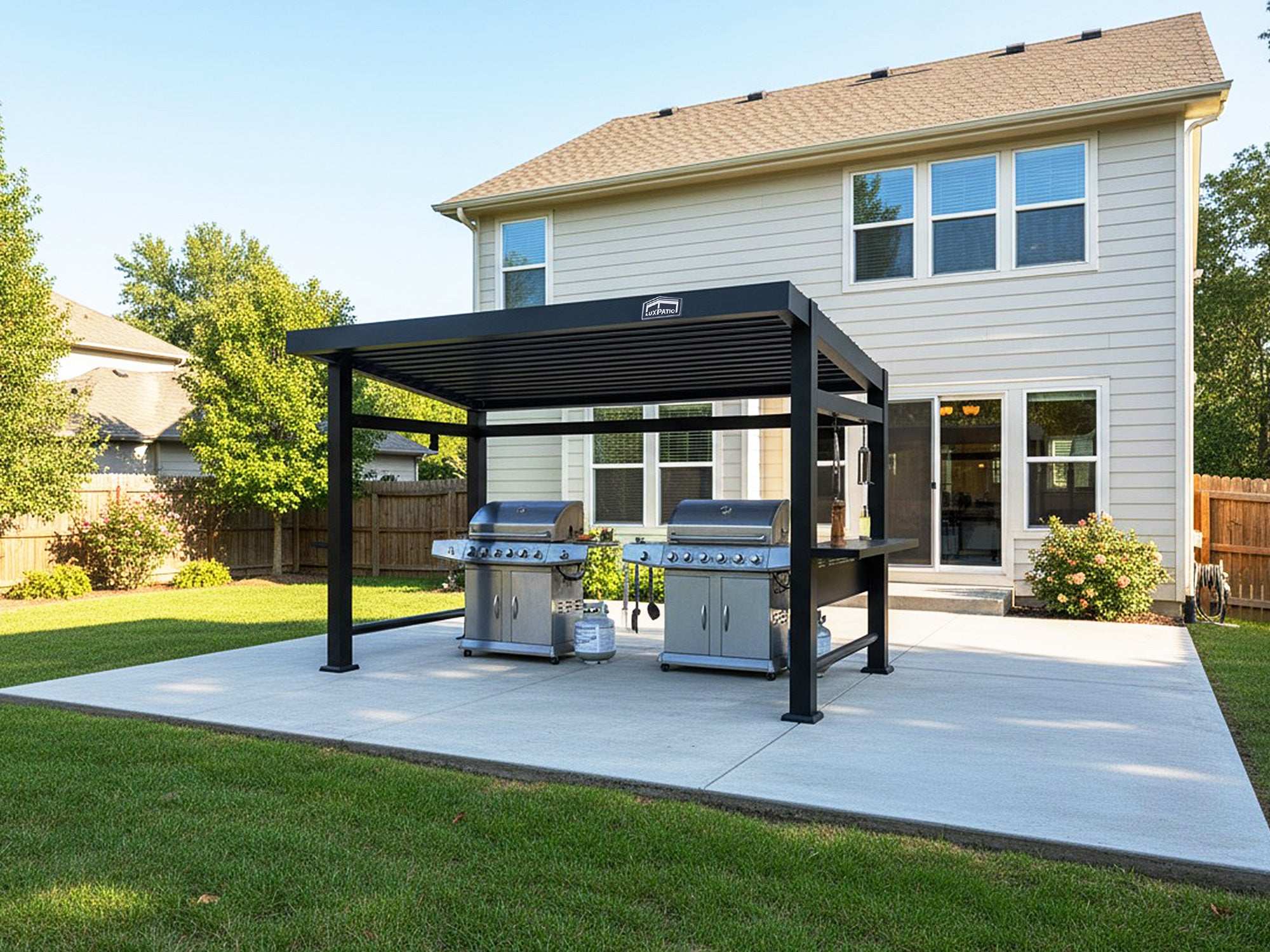 A black pergola with a louvered roof covering two large silver grills on a concrete patio behind a house.